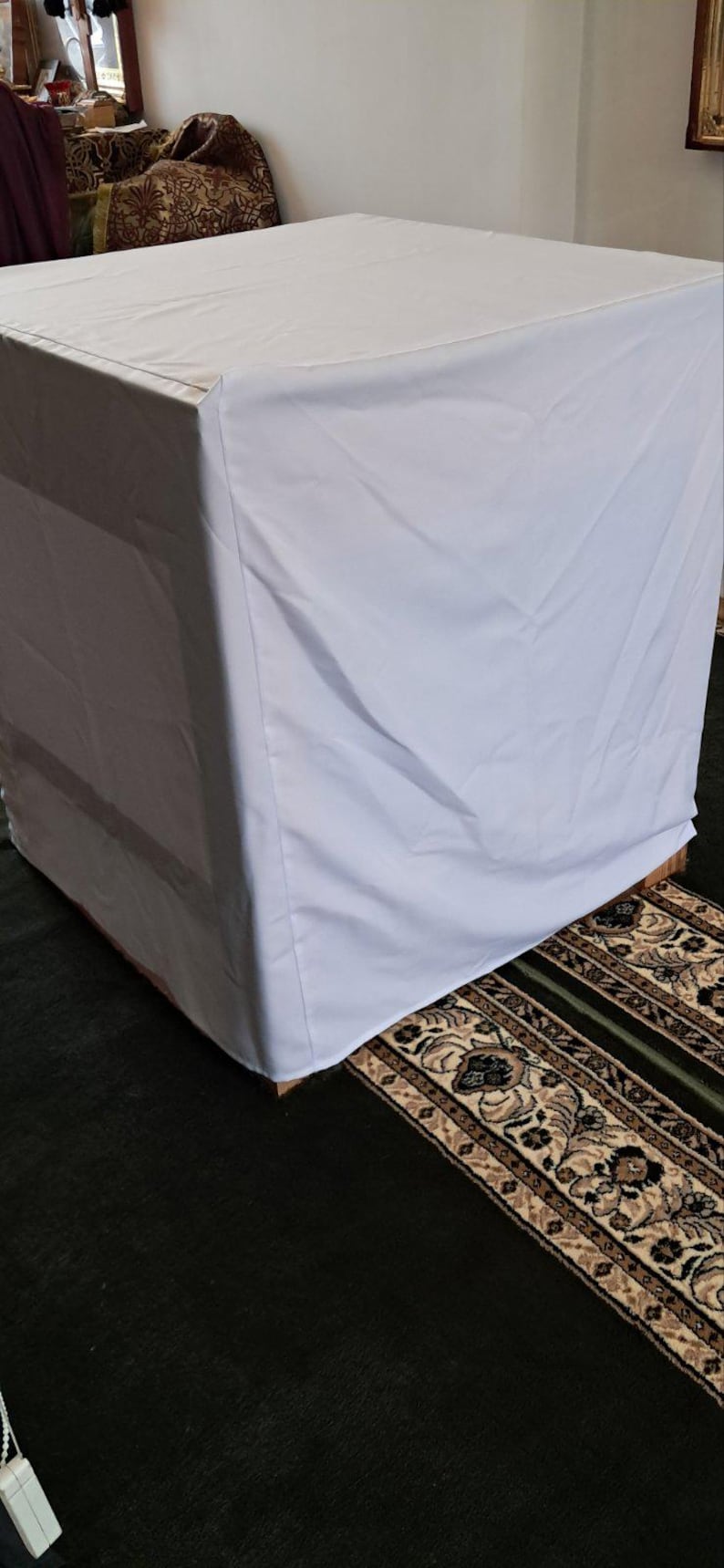 Op de afbeelding: Een witte tafelhoes over een vierkante tafel. De tafel staat op een patroonkleed met een donkergroene achtergrond. De stof lijkt glad en schoon.