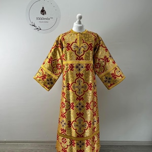May include: A gold-colored liturgical vestment with a red cross pattern. The garment has long sleeves and a high neckline. The text "Ekklesia Church clothes" is visible on a circular logo.