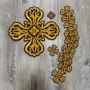 May include: A collection of embroidered patches in gold and brown. The set includes a large ornate cross, two smaller crosses, and a chain of geometric cross shapes. The patches are likely for decorative purposes.