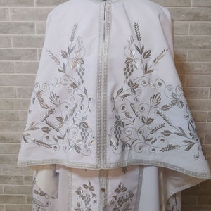 May include: White liturgical vestment with silver embroidery of grapevines and leaves. The vestment has a long, flowing cape and a shorter, fitted undergarment.