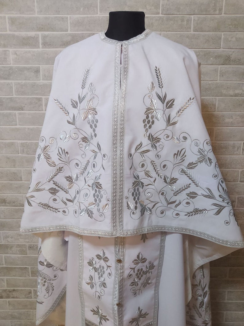 May include: White liturgical vestment with silver embroidery of grapevines and leaves. The vestment has a long, flowing cape and a shorter, fitted undergarment.