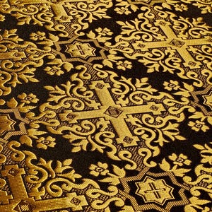 Pu&ograve; includere: Primo piano di un tessuto a fantasia oro e nero. Il disegno presenta ripetuti motivi a croce, floreali e volute ornate. Il tessuto ha un aspetto strutturato, con una leggera lucentezza.