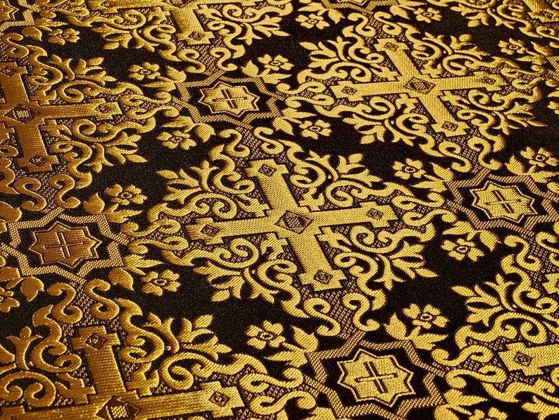 Pu&ograve; includere: Primo piano di un tessuto a fantasia oro e nero. Il disegno presenta ripetuti motivi a croce, floreali e volute ornate. Il tessuto ha un aspetto strutturato, con una leggera lucentezza.