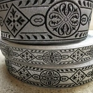 Puede incluir: Dos rollos de ribete plateado y negro con estampado. El ribete tiene un dise&ntilde;o repetitivo de forma de diamante con una cruz en el centro y un c&iacute;rculo con un dise&ntilde;o floral.