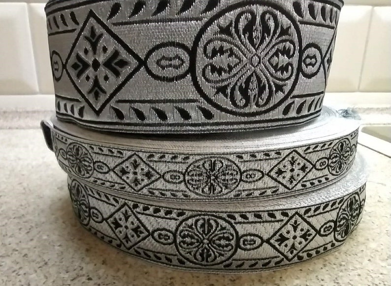 Puede incluir: Dos rollos de ribete plateado y negro con estampado. El ribete tiene un dise&ntilde;o repetitivo de forma de diamante con una cruz en el centro y un c&iacute;rculo con un dise&ntilde;o floral.