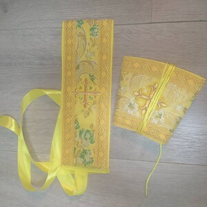 May include: A yellow fabric sash with a gold embroidered cross and floral pattern. The sash is tied with a yellow ribbon. A matching yellow fabric pouch with a gold embroidered cross and floral pattern is also included.