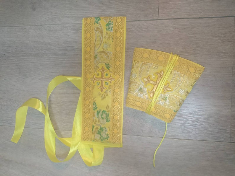 May include: A yellow fabric sash with a gold embroidered cross and floral pattern. The sash is tied with a yellow ribbon. A matching yellow fabric pouch with a gold embroidered cross and floral pattern is also included.