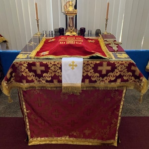 Op de afbeelding: Een rood en gouden altaar doek met een wit kruis en gouden franjes. Het doek ligt op een tafel met een rood fluwelen kussen en een gouden kruis erop. De tafel is versierd met kaarsen en andere religieuze voorwerpen.