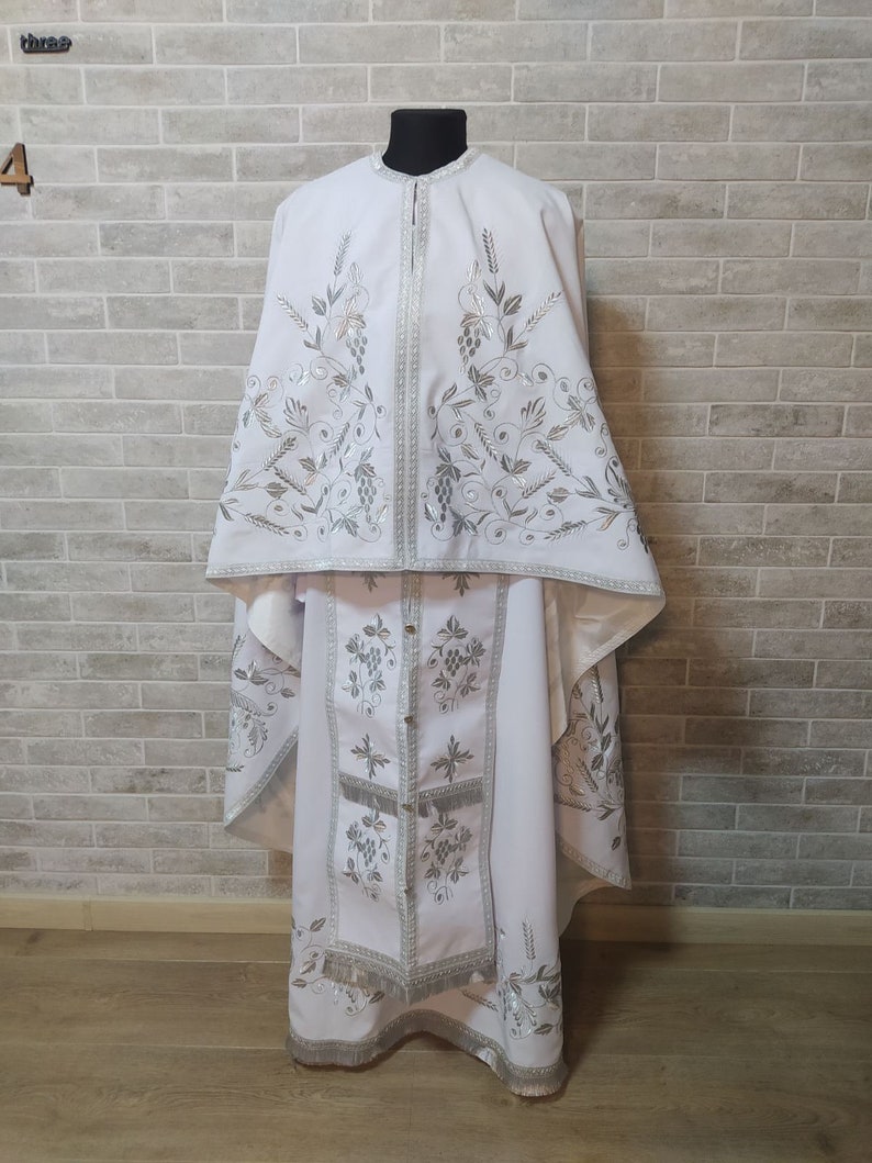May include: White liturgical vestments with silver embroidery of grapevines and wheat. The vestments include a long robe, a shorter robe, and a stole.