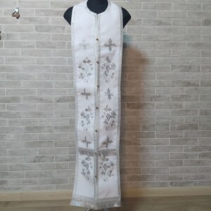 May include: White liturgical vestment with silver embroidery of grapes and crosses. The vestment is displayed on a stand.
