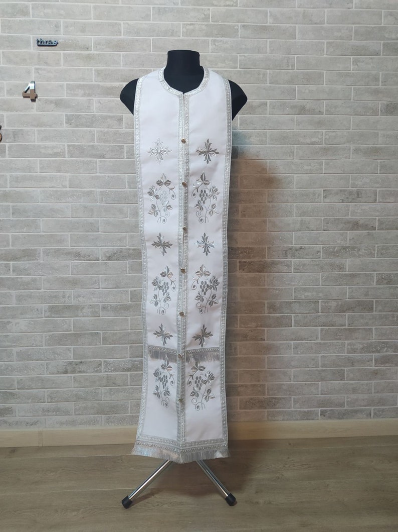 May include: White liturgical vestment with silver embroidery of grapes and crosses. The vestment is displayed on a stand.
