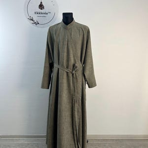 May include: A long, olive-green robe with a mandarin collar and long sleeves, tied at the waist with a matching fabric belt. The robe is displayed on a black mannequin. The logo for Ekklesia Church Clothes is visible in the background.