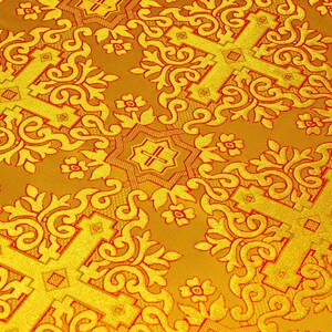 Pu&ograve; includere: Primo piano di un tessuto giallo dorato con un motivo ripetuto di croci e disegni floreali in rosso a contrasto. Il tessuto ha un aspetto strutturato, con il motivo leggermente in rilievo.