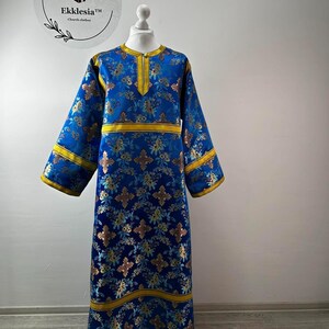 May include: A long, blue liturgical garment with gold accents and floral patterns. The garment features long sleeves and is displayed on a mannequin. The garment has the text "Ekklesia Church clothes" on a circular logo.