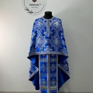 May include: A blue and silver liturgical vestment with intricate floral patterns. The vestment has a long, flowing design and is adorned with silver trim. The text "Ekklesia™ Church clothes" is visible in the top left corner of the image.