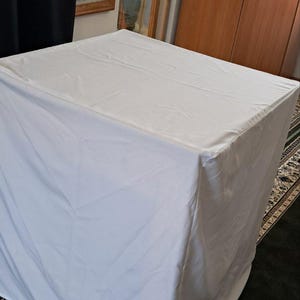 Op de afbeelding: Een witte rechthoekige tafel bedekt met een witte doek. De tafel staat in een kamer met religieuze kunstwerken aan de muur en een patroonkleed op de vloer. De tafel wordt waarschijnlijk gebruikt voor religieuze doeleinden.