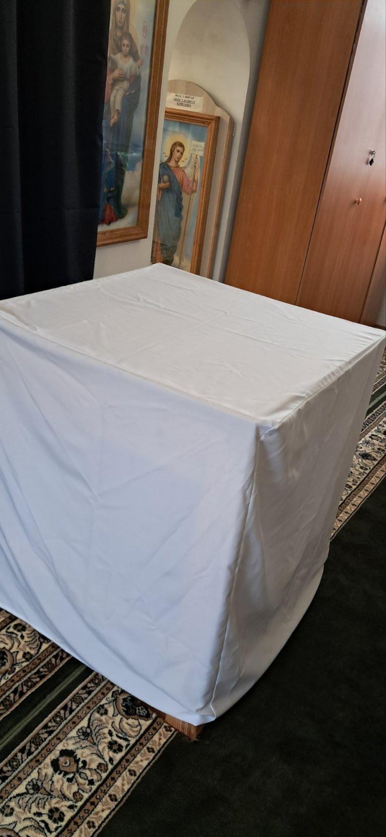 Op de afbeelding: Een witte rechthoekige tafel bedekt met een witte doek. De tafel staat in een kamer met religieuze kunstwerken aan de muur en een patroonkleed op de vloer. De tafel wordt waarschijnlijk gebruikt voor religieuze doeleinden.