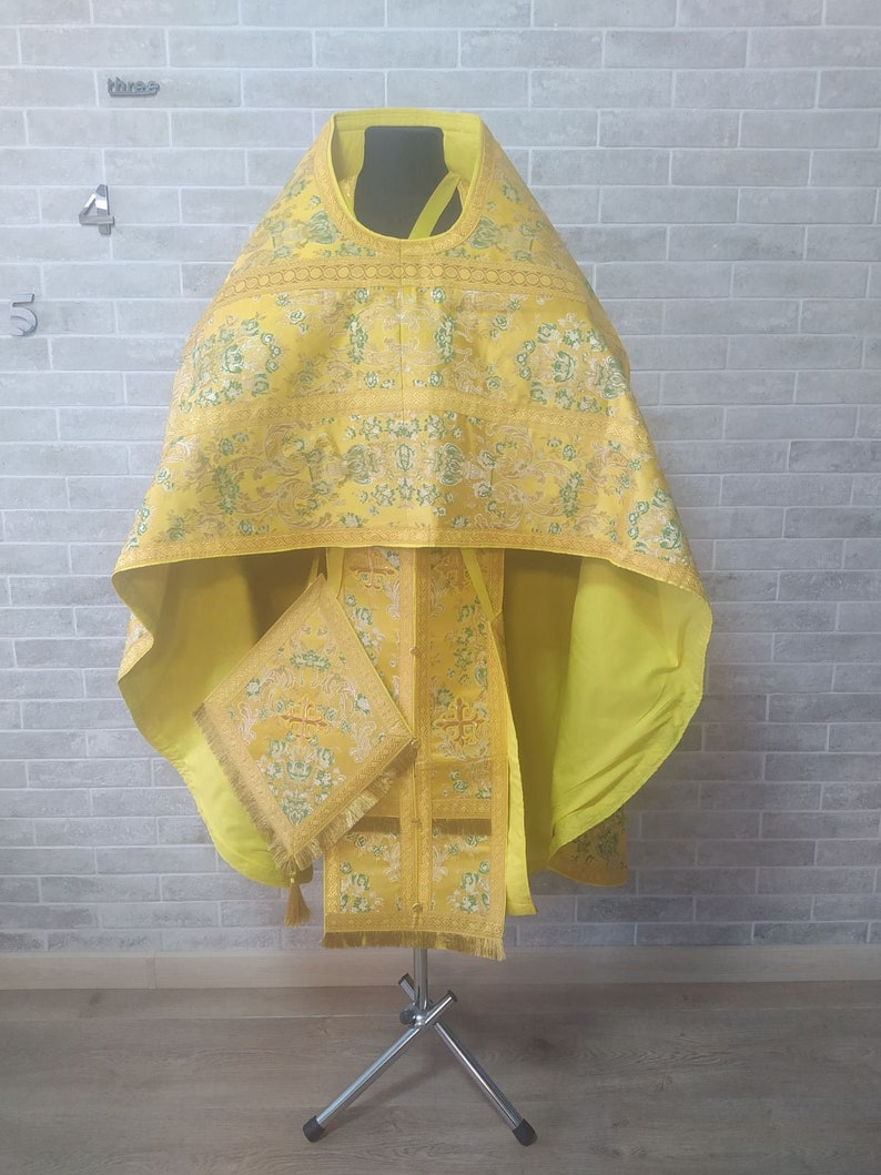 May include: A yellow liturgical vestment with gold embroidery and a matching stole. The vestment is displayed on a tripod stand against a brick wall.