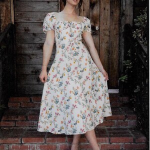 May include: A white floral print dress with a square neckline and short puff sleeves. The dress features a colorful floral pattern and a knee-length skirt. The dress is worn with clear sandals. The brand name "Brandi Joan" is visible in the bottom right corner.