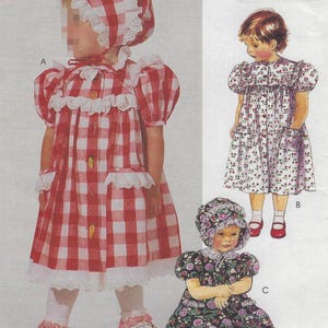 May include: A vintage sewing pattern featuring three children's dresses. The dresses include a red and white gingham dress with a matching bonnet, a floral print dress, and a dark floral dress with a bonnet. The pattern includes detailed illustrations.