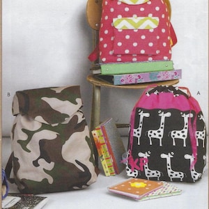 May include: Three different backpacks, one with a camouflage pattern, one with a pink polka dot pattern and a green chevron flap, and one with a black background and white giraffe print. The backpacks are on a wooden chair and a table with books and notebooks.