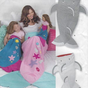 May include: Mermaid tail blankets in pink, blue, and teal with star and flower appliqués. A gray shark-shaped sleeping bag is also shown. The blankets are designed for children and dolls. The image shows a woman and child.