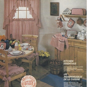 May include: A kitchen decorated in a country style with red and white gingham check curtains, tablecloths, and chair cushions. The kitchen features a wooden table with four chairs, a wooden cabinet, and a shelf with various kitchen accessories. The text "Kitchen Essentials" and "Les Indispensables de la Cuisine" is visible in the image.