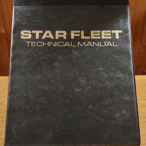 May include: A black Star Fleet Technical Manual with gold lettering. The manual is in a binder and appears to be vintage. The cover is slightly worn, suggesting it has been used.