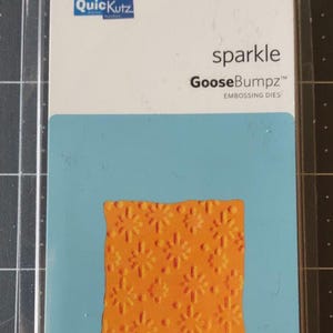 May include: A QuickKutz GooseBumpz embossing die package. The package features a blue and white card with the word "sparkle" and the product name. The die itself is orange and has a floral pattern. The package includes one embossing die.