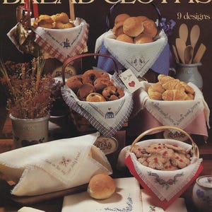May include: A collection of nine cross-stitch bread cloth patterns featuring various designs, including "Give us this day our daily bread", "Home sweet home", and "Tea". The patterns are displayed on baskets filled with bread, rolls, and cookies.