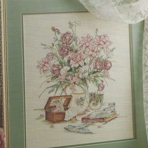 May include: A framed cross-stitch embroidery depicting a bouquet of pink and mauve roses in a white pitcher, accompanied by an open wooden box, a small bowl, a closed book, and a pair of scissors, all set against a light cream background.