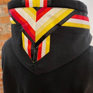 May include: Black hooded sweatshirt with a colorful geometric pattern on the hood. The pattern is made of red, yellow, white, and black ribbon.