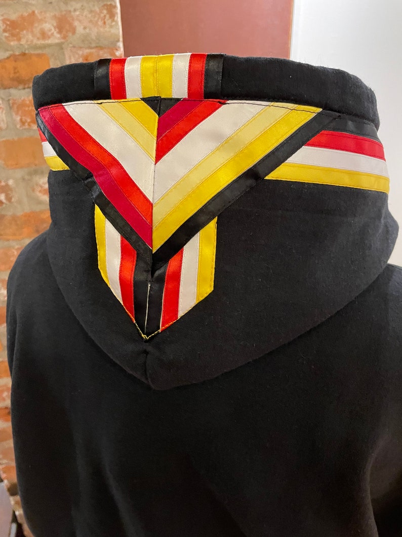 May include: Black hooded sweatshirt with a colorful geometric pattern on the hood. The pattern is made of red, yellow, white, and black ribbon.