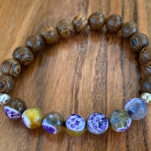 May include: A beaded bracelet featuring alternating wooden and gemstone beads. The wooden beads have a dark brown, striped pattern. The gemstone beads are purple, yellow, and gray with unique patterns. Silver-colored beads separate the two bead types.