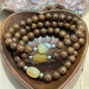 May include: Two brown wooden beaded bracelets with three yellow and white gemstone beads. The bracelets are displayed in a heart-shaped wooden bowl.