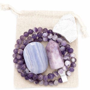 May include: A collection of crystals and beads in a small beige drawstring bag. The set includes a blue lace agate stone, a purple crystal point, a white stone, and a beaded necklace with purple beads.