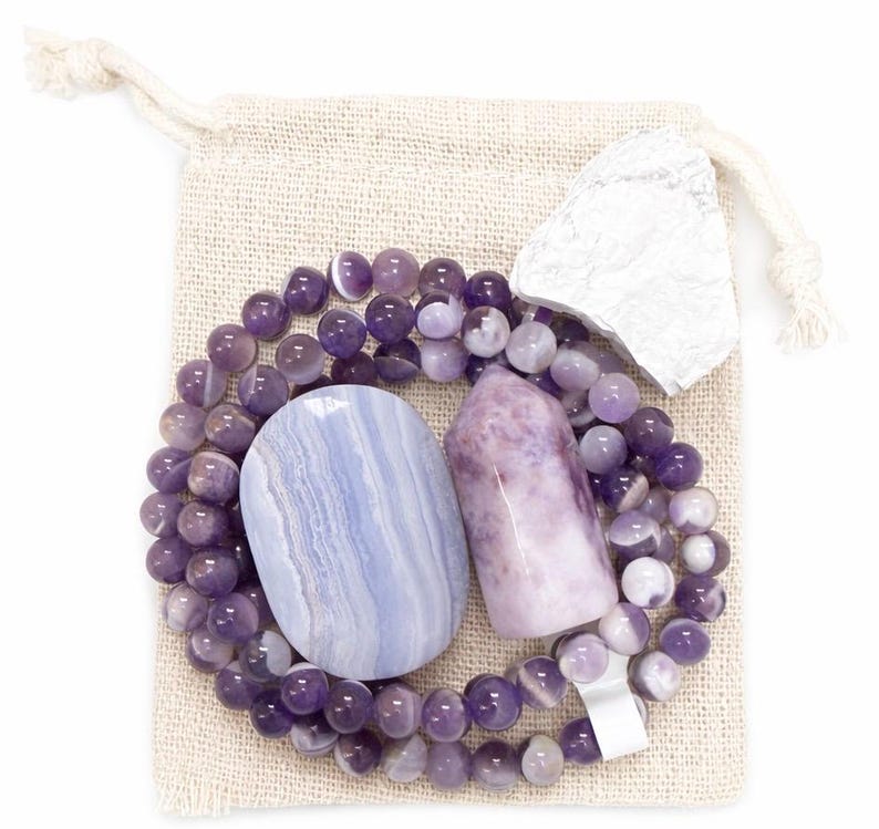 May include: A collection of crystals and beads in a small beige drawstring bag. The set includes a blue lace agate stone, a purple crystal point, a white stone, and a beaded necklace with purple beads.
