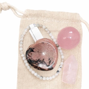 May include: A collection of crystals and a beaded bracelet are displayed inside a beige drawstring bag. The crystals include a pink heart-shaped stone, a sphere, and a pointed obelisk. The bracelet is made of small, round, light-colored beads.