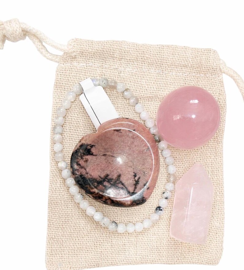 May include: A collection of crystals and a beaded bracelet are displayed inside a beige drawstring bag. The crystals include a pink heart-shaped stone, a sphere, and a pointed obelisk. The bracelet is made of small, round, light-colored beads.