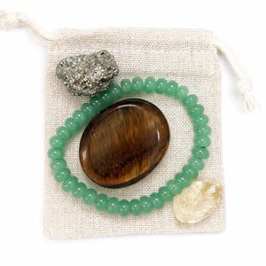 May include: A collection of crystals and a beaded bracelet inside a small, beige drawstring bag. The crystals include a pyrite cluster, a brown oval stone, and a pale yellow stone. The bracelet is made of small, round, green beads.