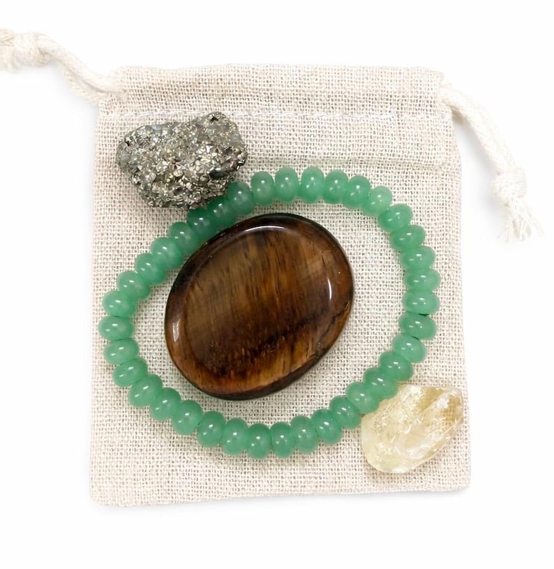 May include: A collection of crystals and a beaded bracelet inside a small, beige drawstring bag. The crystals include a pyrite cluster, a brown oval stone, and a pale yellow stone. The bracelet is made of small, round, green beads.