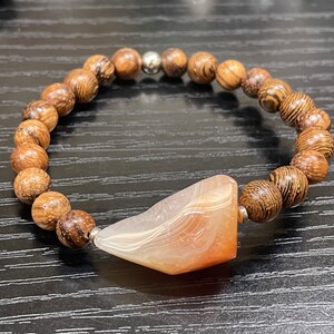 May include: A brown wooden beaded bracelet with a light brown agate pendant. The pendant is shaped like a teardrop and has white and orange stripes.
