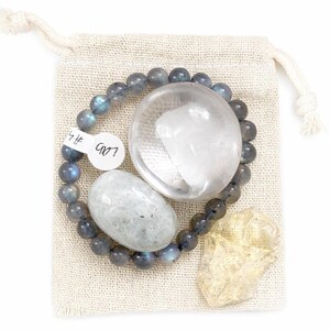 May include: A collection of crystals and a beaded bracelet arranged on a small beige drawstring bag. The bracelet features gray beads. A large clear crystal, a gray oval stone, and a yellow crystal are also included.