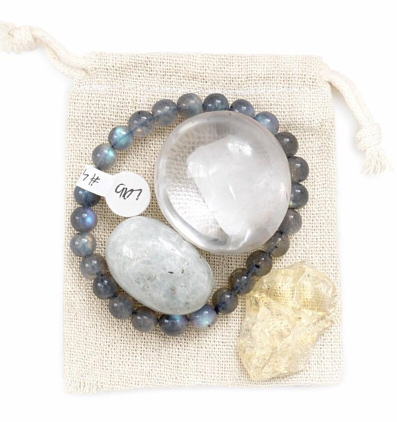 May include: A collection of crystals and a beaded bracelet arranged on a small beige drawstring bag. The bracelet features gray beads. A large clear crystal, a gray oval stone, and a yellow crystal are also included.