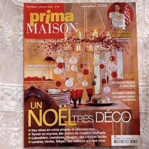 Op de afbeelding: Een tijdschriftcover met een kerstthema met een kroonluchter met kaarsen en ornamenten, een gedekte tafel met rode wijnglazen en de tekst "prima MAISON" en "UN NOEL TRES DECO".