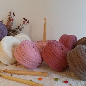Può includere: Una collezione di gomitoli di lana colorati in tonalità di viola, rosa, bianco e marrone. La lana è disposta su una superficie bianca con due uncinetti di legno e piccoli oggetti decorativi.