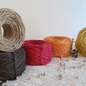 Op de afbeelding: Vijf spoelen garen in verschillende kleuren, waaronder naturel, bruin, rood, oranje en geel. De spoelen zijn gerangschikt op een wit oppervlak met zilveren kralen eromheen.