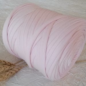 Puede incluir: Una gran bola cilíndrica de hilo de tela rosa claro. El hilo está bien enrollado, con líneas paralelas visibles. El fondo es una superficie texturizada de color blanquecino. Unas espigas de trigo están en la esquina inferior izquierda.