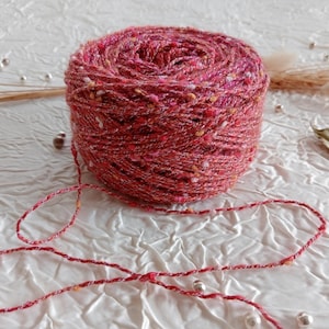 Op de afbeelding: Een bol rood garen met witte en gouden vlekken. Het garen ligt op een wit oppervlak met een paar kleine witte kralen eromheen.