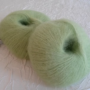 May include: Two skeins of light green yarn, likely mohair or a similar soft fiber, are shown on a white textured surface. The yarn is wound into balls and ready for knitting or crocheting.